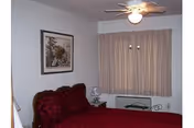 A bedroom with a red bedspread on a bed with a decorative headboard, a nightstand with a lamp, a framed picture on the wall, beige curtains covering a window, and a ceiling fan with a light.