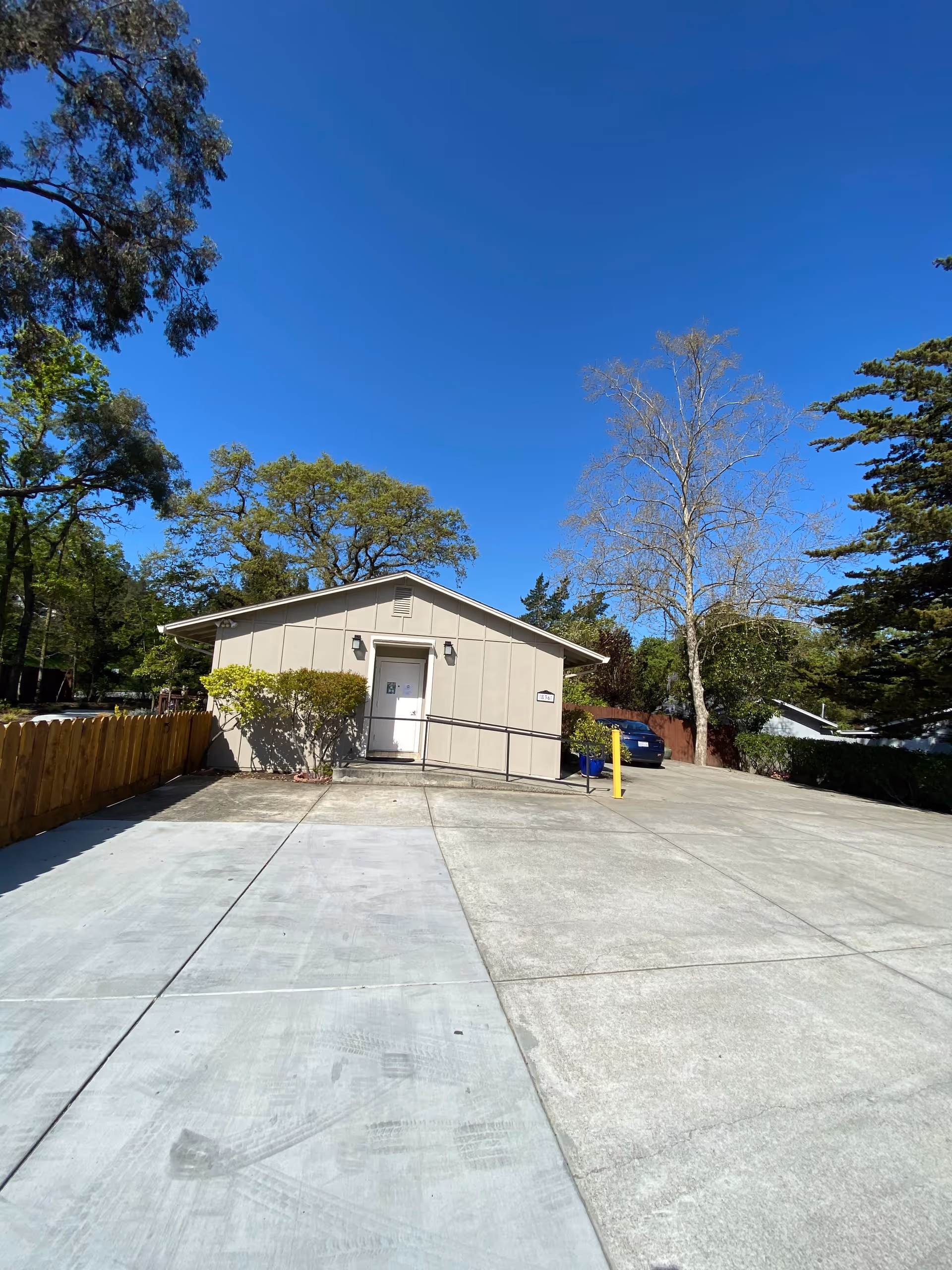 Small single-story building with a light beige exterior, a white door with a ramp leading up to it, surrounded by trees and bushes under a clear blue sky. There is a concrete driveway and parking area in front of the building.