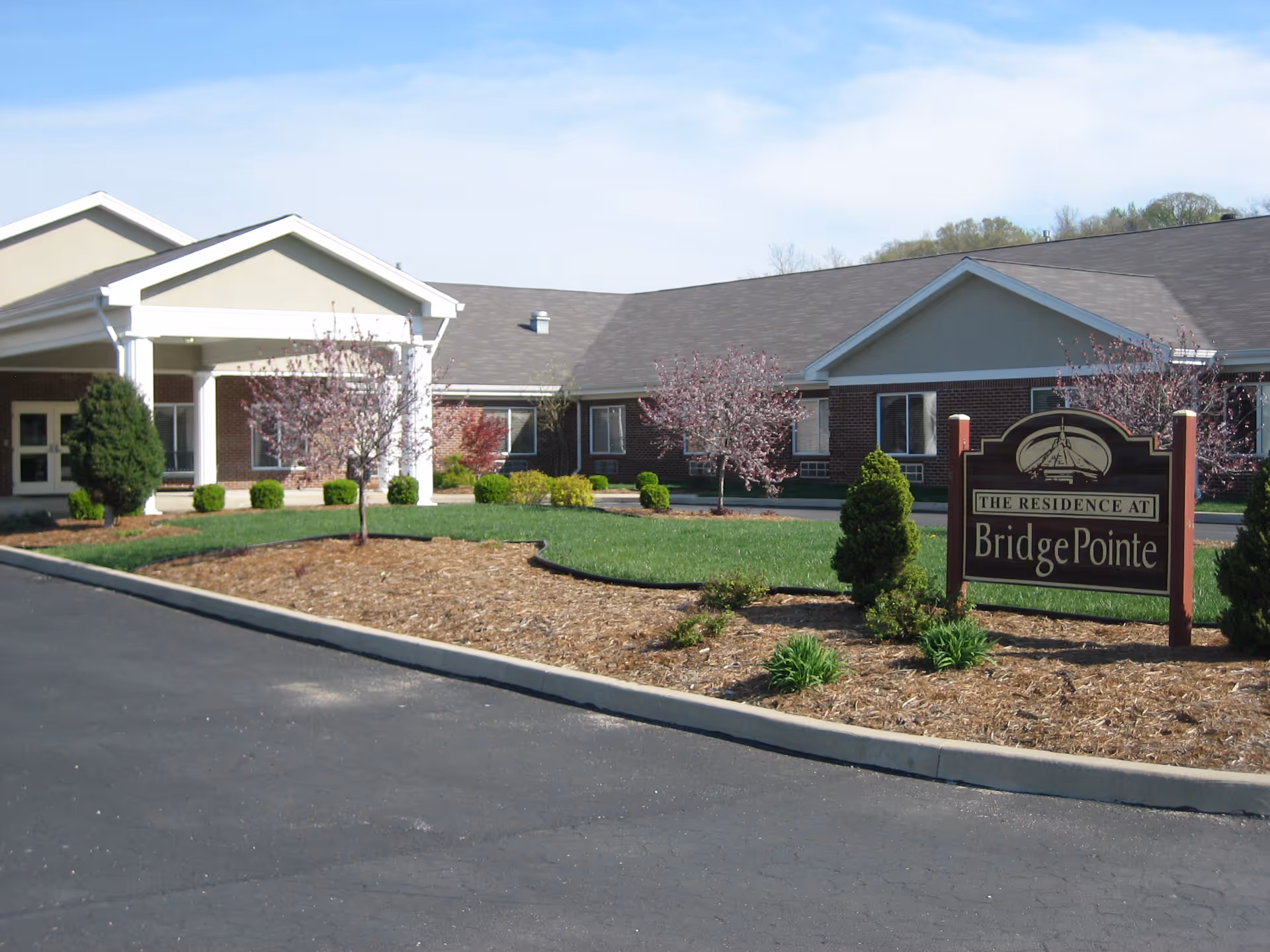 Exterior view of a single-story brick building with a covered entrance, landscaped with small trees and bushes, and a sign in front that reads 'The Residence at BridgePointe'.