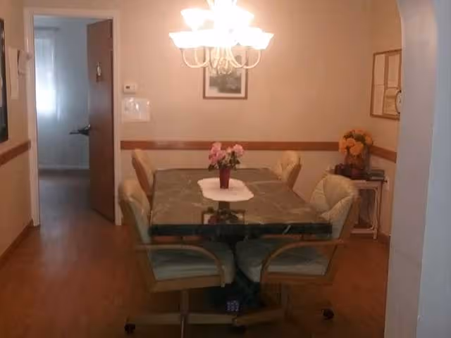 A dining room with a rectangular table covered by a green tablecloth and a white runner, surrounded by six cushioned chairs on wheels. A chandelier with multiple lights hangs above the table. There is a small vase with pink flowers on the table. The room has wooden flooring and beige walls with a wooden chair rail. A doorway leads to another room with a window letting in natural light. On the right side, there is a small table with a bouquet of orange flowers and some framed pictures on the wall.