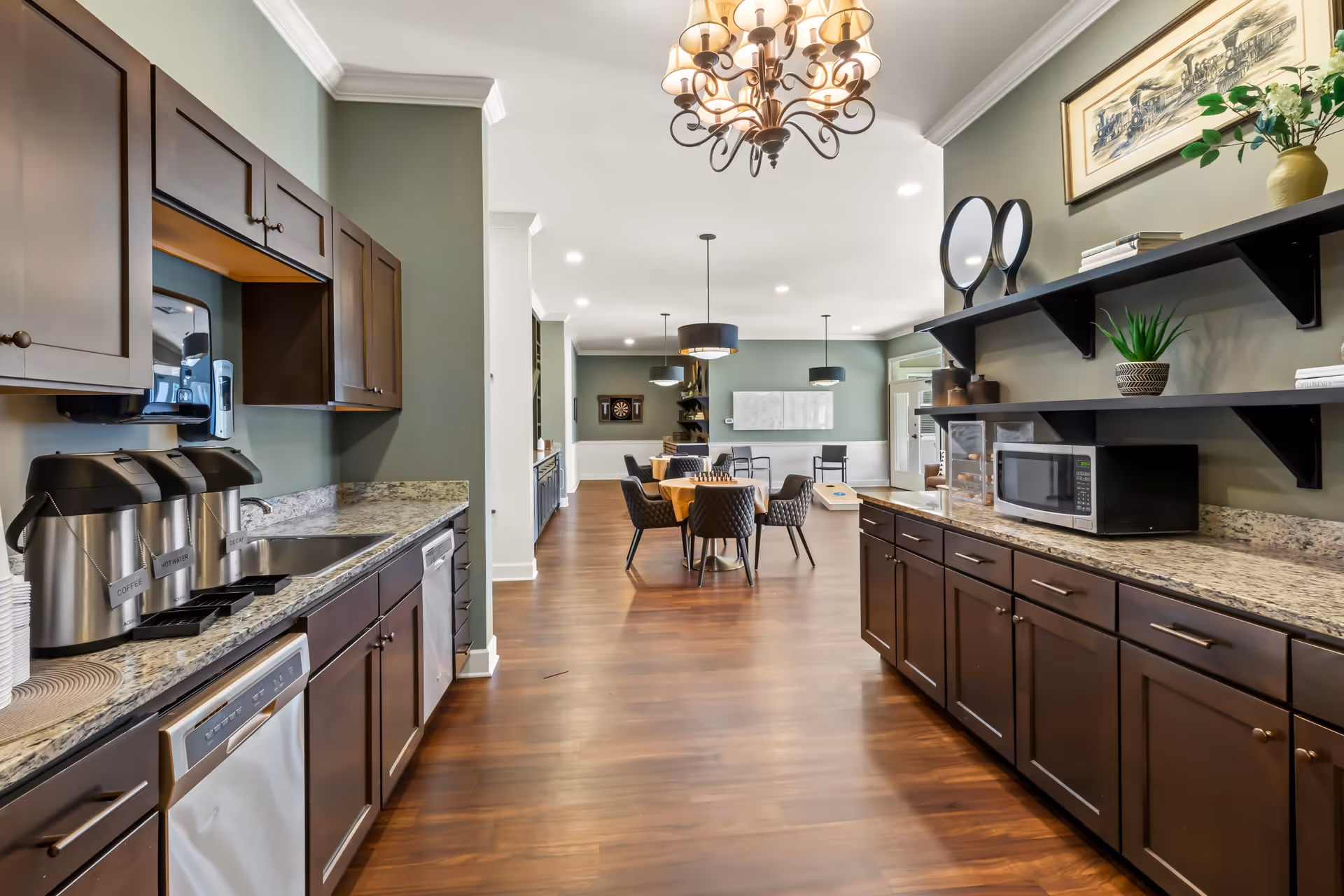 A spacious kitchen area with dark wood cabinets, granite countertops, and stainless steel appliances including a dishwasher and coffee dispensers. The room opens into a dining area with a table and chairs, modern pendant lights, and a dartboard on the far wall. Shelves on the right hold decorative items and a microwave. The floor is wooden, and the walls are painted a muted green.