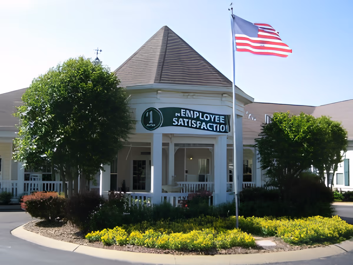 Exterior view of a senior living facility building with a peaked roof, white columns, and a porch area. There is a flagpole with an American flag in front of the building, surrounded by green bushes and yellow flowers. A sign on the building reads '#1 Employee Satisfaction'.