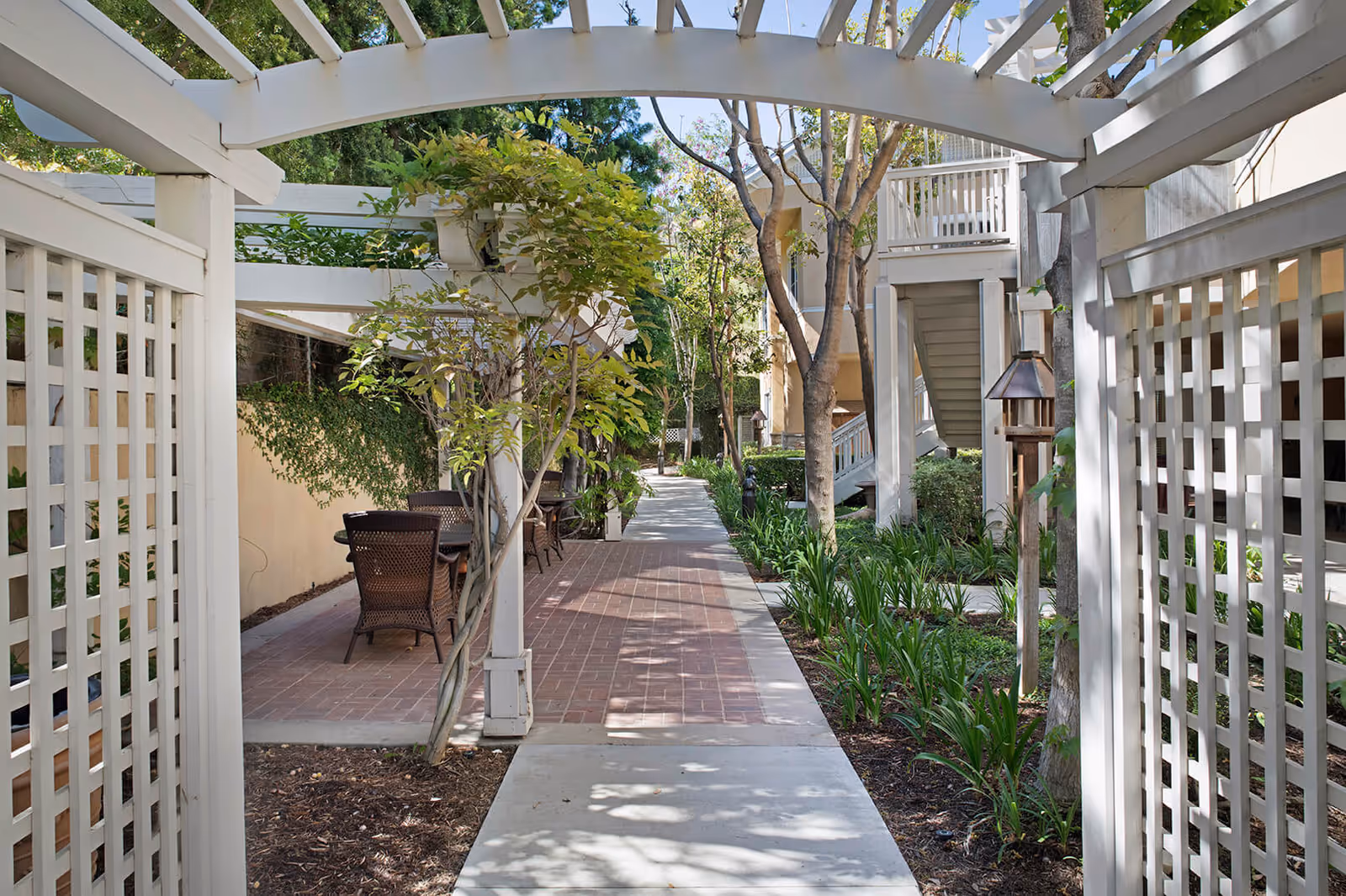 A shaded outdoor walkway at a senior living facility with white wooden pergolas, green plants, trees, and outdoor seating with wicker chairs and tables along the side.