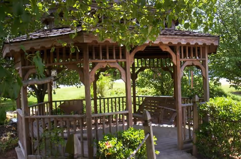 A wooden gazebo surrounded by green trees and bushes in an outdoor garden area, with a wooden bench swing inside the gazebo.