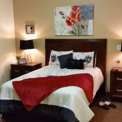 A cozy bedroom with a neatly made bed featuring white and floral bedding, a red throw blanket, and decorative pillows. There are two dark wooden nightstands on either side of the bed, each with a lamp. A small plant and a framed photo are on the left nightstand. A floral painting hangs on the wall above the bed. A pair of slippers is placed on the floor near the right nightstand.