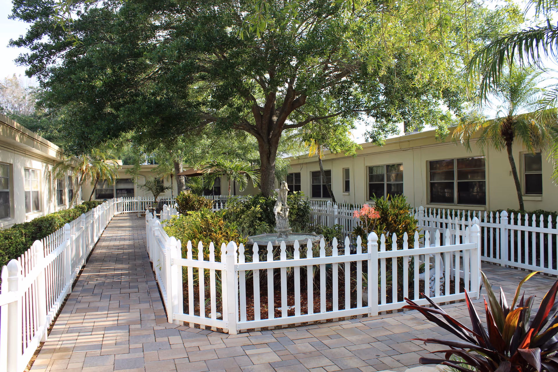 Outdoor courtyard area at Apollo Health and Rehabilitation Center featuring a paved walkway lined with white picket fences, a central garden with various plants and a tree, and a small statue or fountain in the middle surrounded by greenery.