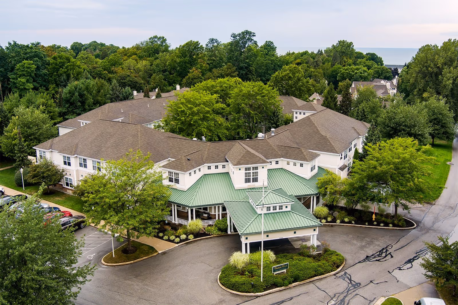 Aerial view of Westlake Woods Assisted Living facility surrounded by trees and greenery, showing a large building with a green roof entrance and a parking area with several cars.