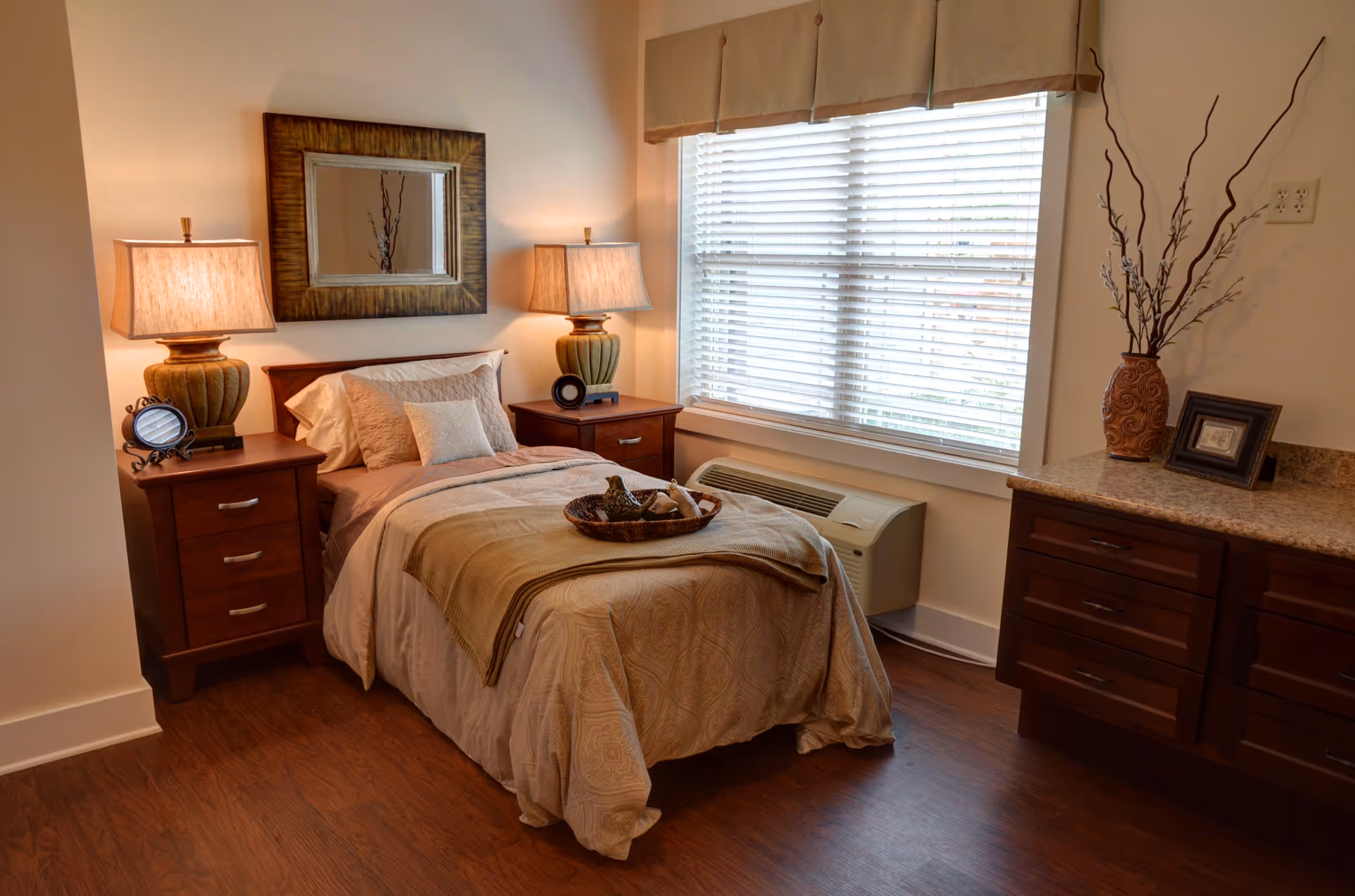A cozy bedroom with a single bed covered in beige bedding and a decorative tray on top. There are two wooden nightstands on either side of the bed, each with a lamp and small decorative items. A large window with white blinds lets in natural light. A framed mirror hangs above the bed, and a wooden dresser with a vase and picture frame is visible on the right side of the room. The floor is wooden.