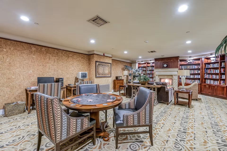 A cozy common room with a round card table and four patterned chairs in the foreground. The room features a fireplace surrounded by built-in wooden bookshelves filled with books. There are additional seating areas with sofas and armchairs, decorative plants, and warm lighting from ceiling fixtures and table lamps.