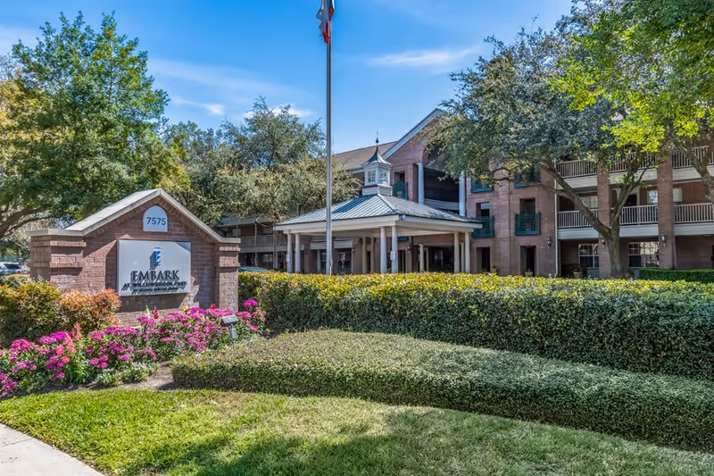 Exterior front entrance of a brick senior living building with a covered porte-cochere, flagpole, landscaping, and a sign at the driveway.