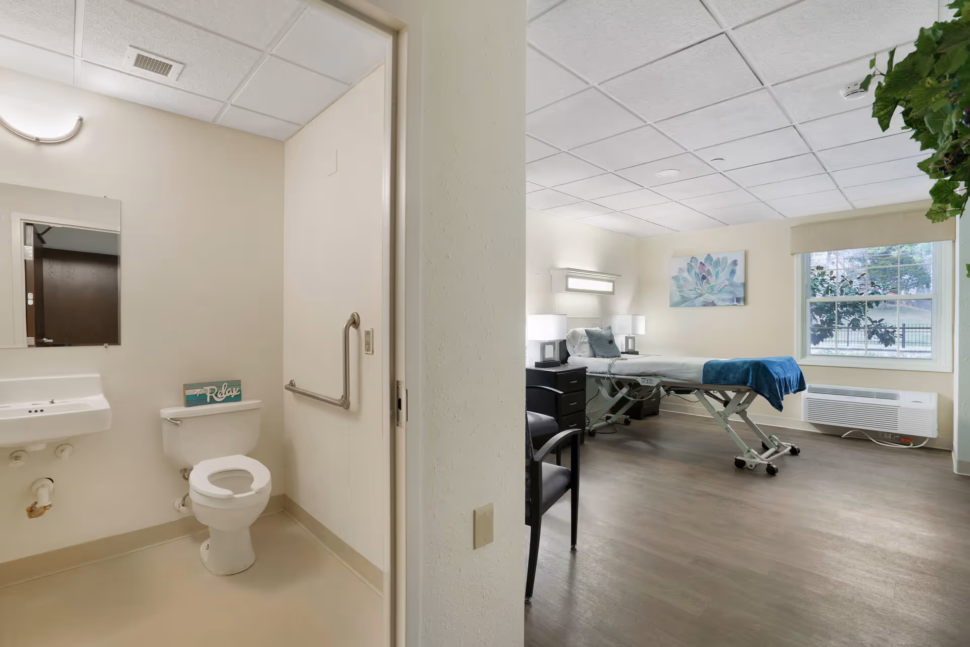 View of a senior living facility room showing a bathroom on the left with a toilet, sink, mirror, and a small sign that says 'Relax'. On the right is a bedroom area with a hospital-style bed, a chair, a nightstand with lamps, a large window with a view of greenery outside, and a wall-mounted air conditioning unit.