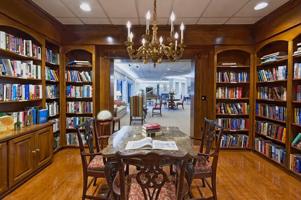 Wood-paneled library room with bookshelves, a marble-topped table and chairs beneath a chandelier and an open view into a seating area.