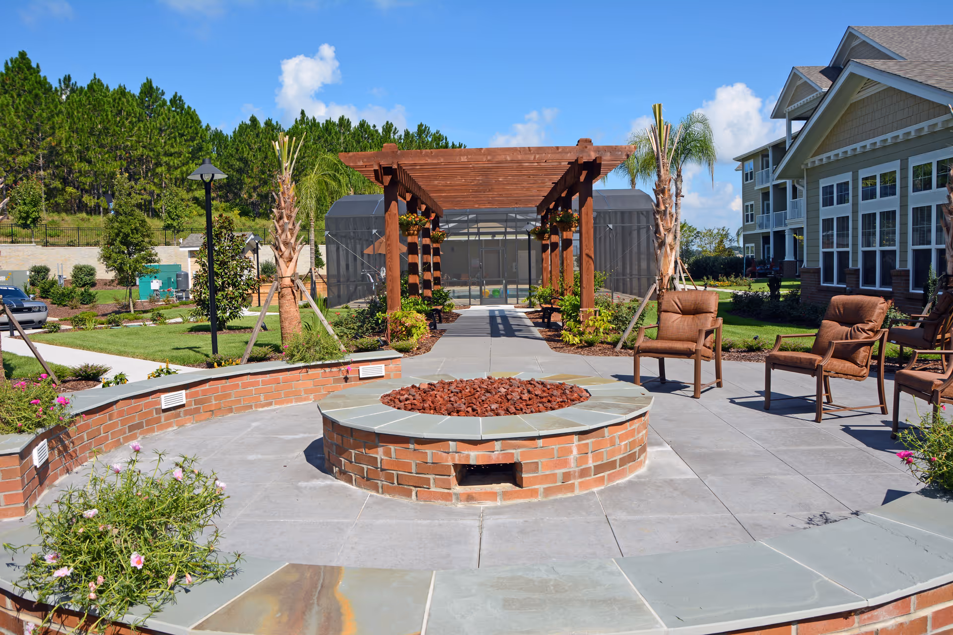 Outdoor patio area at Orchard Heights Gracious Retirement Living featuring a circular brick fire pit surrounded by a curved brick bench and several cushioned chairs. A wooden pergola with hanging plants is visible in the background, along with palm trees, manicured lawns, and a building with large windows under a clear blue sky.