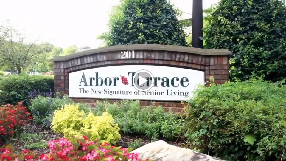 A brick sign surrounded by green bushes and colorful flowers displaying the name 'Arbor Terrace' with the tagline 'The New Signature of Senior Living' and the number 201 above it.
