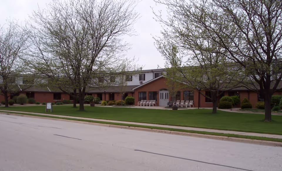 Front view of a two-story brick retirement center with trees, a manicured lawn, and chairs near the entrance.