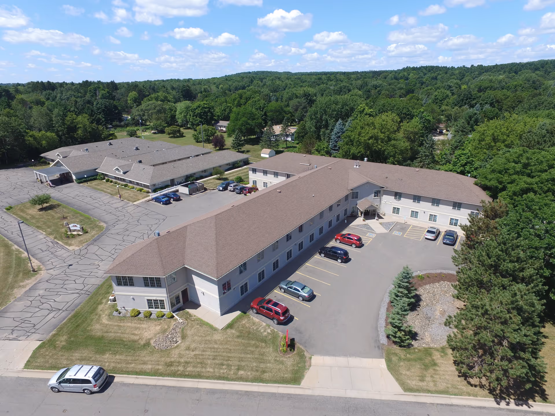 Aerial view of Mountain Terrace Senior Living facility showing a large two-story building with a brown roof surrounded by trees and greenery. Several cars are parked in the parking lot adjacent to the building. The sky is partly cloudy with blue patches.