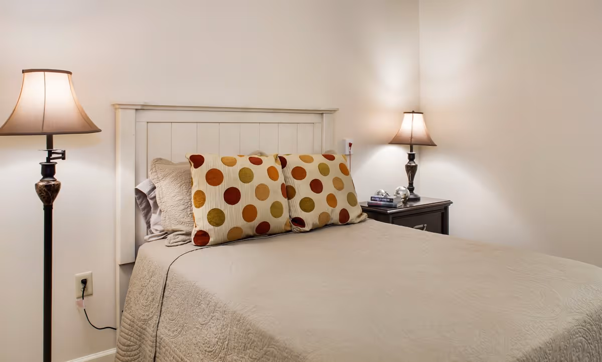 A neatly made bed with a white wooden headboard, beige quilted bedspread, and two large pillows with colorful polka dots. There are two bedside tables, each with a lamp emitting warm light. One bedside table also has a few books and decorative items.