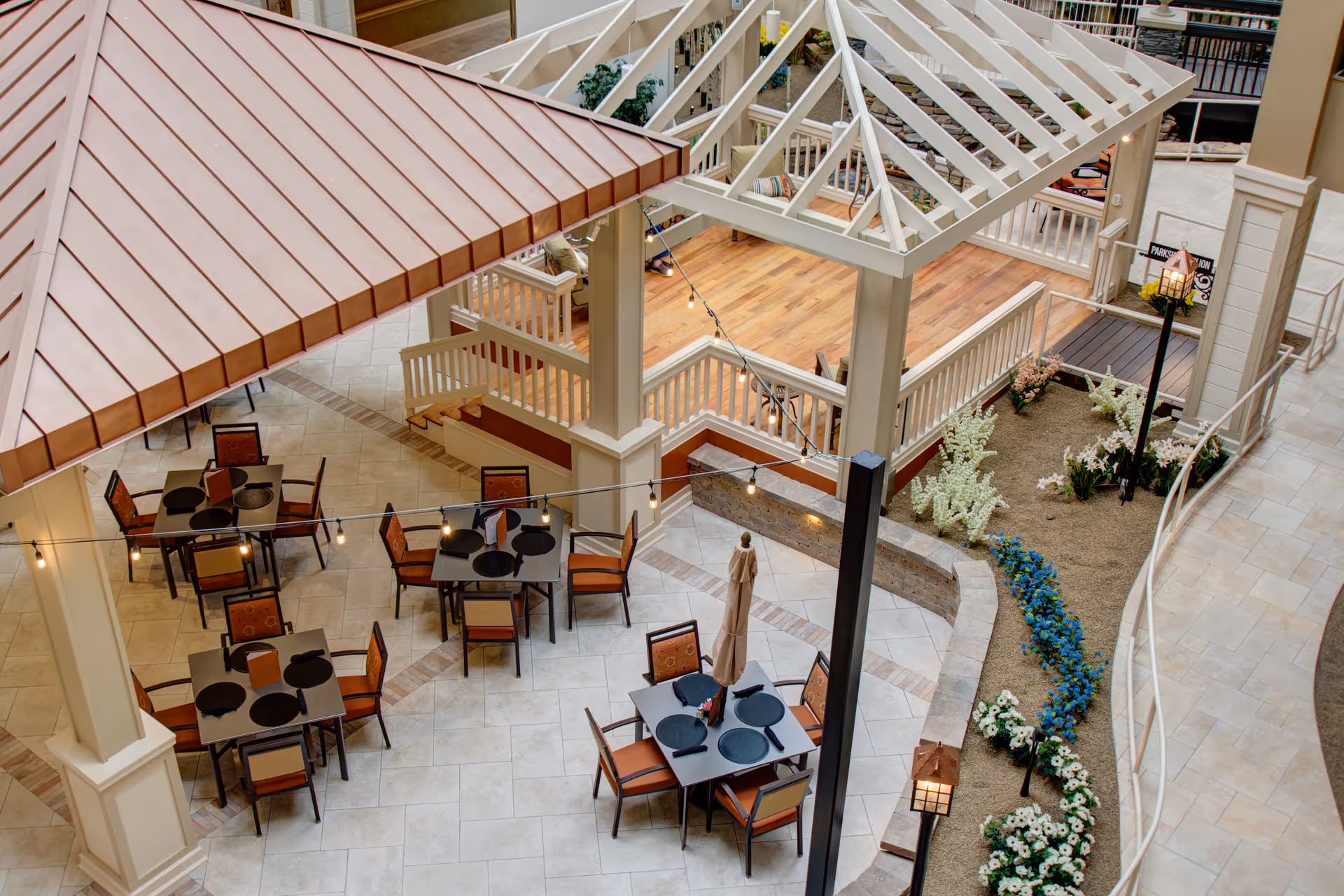 Indoor courtyard area with several square tables and chairs arranged for dining. The space features tiled flooring, a wooden deck area, decorative string lights hanging overhead, and landscaped flower beds with white and blue flowers. There are architectural elements including columns, railings, and a pergola-style roof structure.