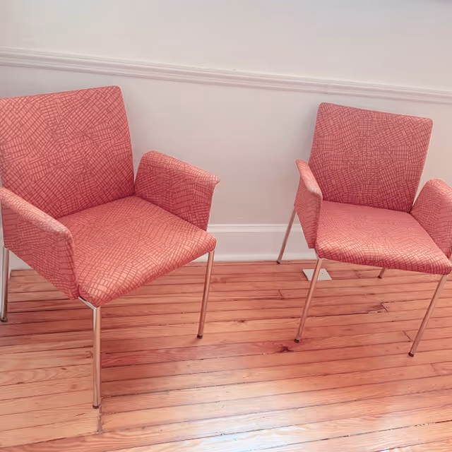 Two pink patterned armchairs with metal legs placed on a wooden floor against a white wall with molding.