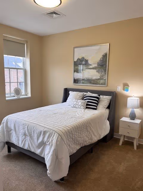 A neatly made bed with white and striped bedding and three decorative pillows in a bedroom with beige walls. There is a window with a roller shade partially down on the left wall, a framed landscape painting above the bed, and a white nightstand with a lamp on the right side of the bed.