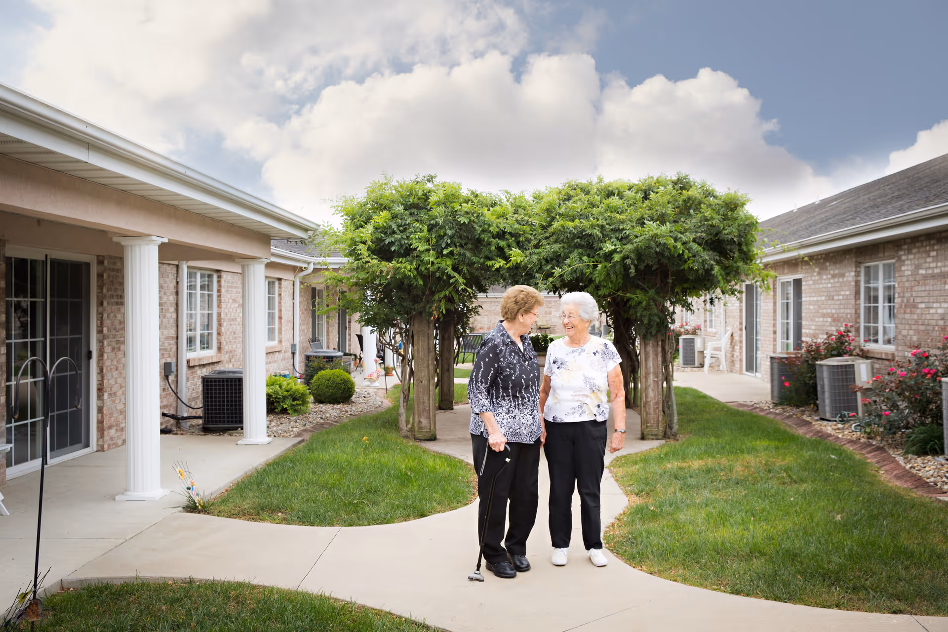 Two elderly women standing and smiling at each other on a paved walkway in a courtyard area of a senior living facility. The courtyard is surrounded by single-story brick buildings with white columns and windows. There is a green archway of foliage behind them and well-maintained grass and shrubs on either side of the walkway.