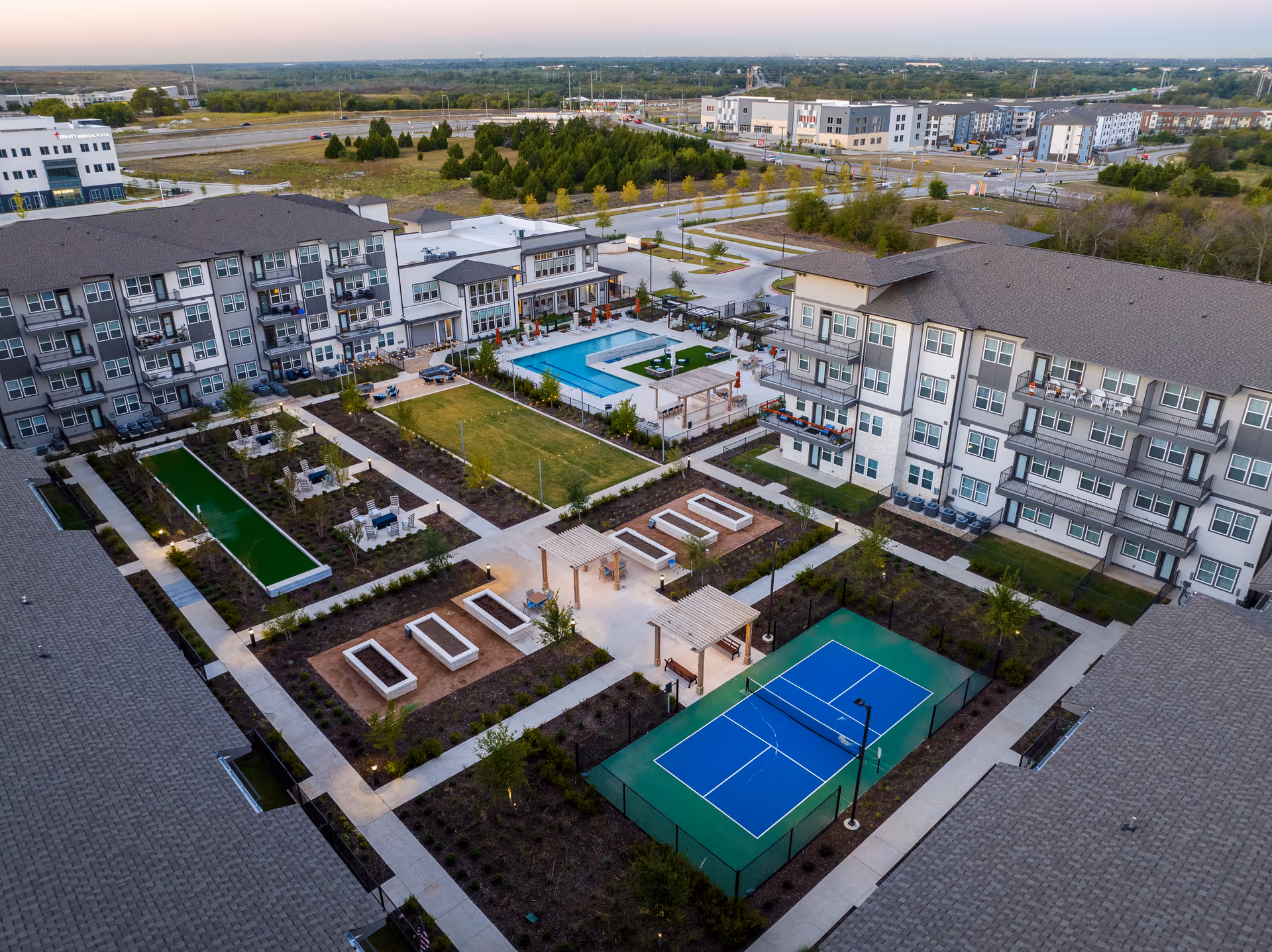 Aerial view of Amberlin at The Station senior living facility showing multiple residential buildings surrounding an outdoor courtyard with a swimming pool, bocce ball courts, a pickleball court, seating areas with pergolas, and landscaped walkways.