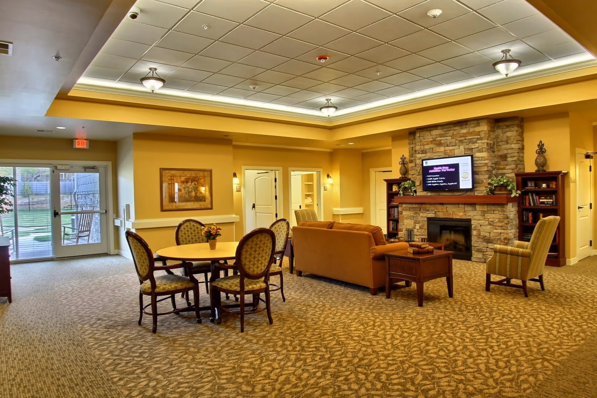 A cozy common area in a senior living facility with a round table and four chairs, a comfortable sofa, an armchair, and a stone fireplace with a mounted TV above it. The room has warm yellow walls, carpeted floors, and a ceiling with recessed lighting. There is a glass door leading outside and framed artwork on the wall.