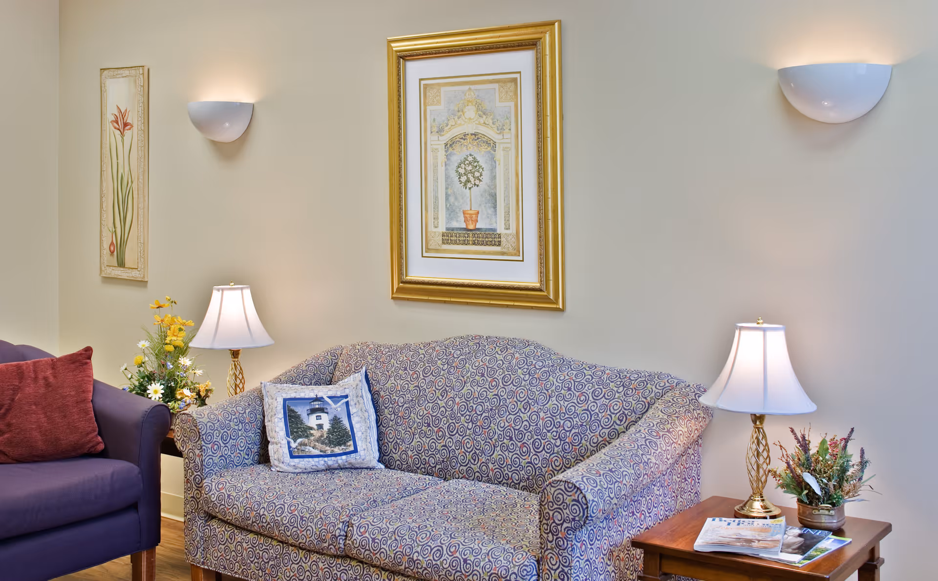 A cozy living room area with a patterned sofa and a purple armchair. The sofa has a decorative pillow with a lighthouse image. There are two table lamps on wooden side tables, one on each side of the sofa, with floral arrangements next to them. The walls are light-colored and decorated with two framed pictures and two wall sconces providing soft lighting.