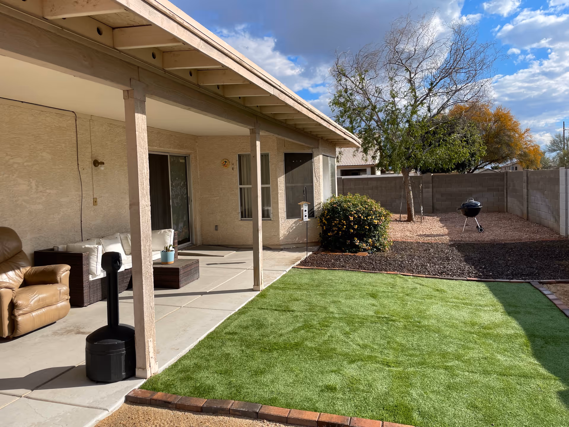 Covered patio with seating beside a small artificial turf lawn, shrub, tree, and a charcoal grill in a fenced backyard.