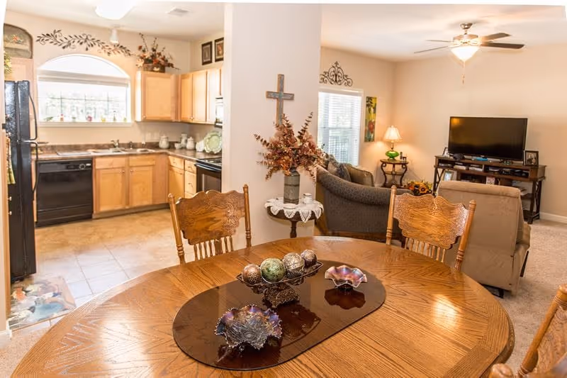 View of a cozy living and dining area in a senior living facility. In the foreground is a wooden dining table with decorative bowls and ornamental balls. Behind the table is a small round side table with a floral arrangement and a wall-mounted cross. To the left is a kitchen with wooden cabinets, a black refrigerator, dishwasher, and stove. To the right is a living room with a ceiling fan, a TV on a stand, a lamp on a side table, and two upholstered chairs.
