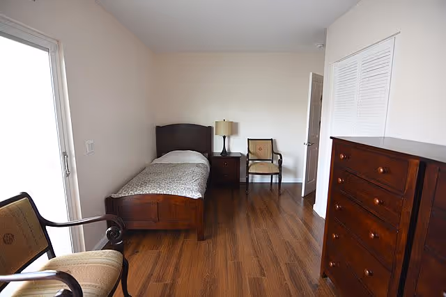 A simple bedroom with a single wooden bed with a patterned bedspread, a wooden nightstand with a lamp, two upholstered chairs, a wooden dresser, and hardwood flooring. The room has white walls and a door leading to another area.