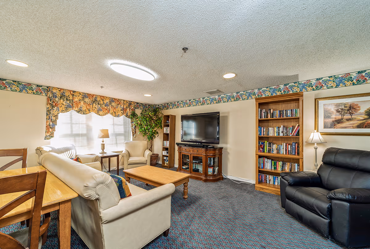 A cozy living room with a beige sofa, an armchair, a wooden coffee table, a black leather recliner, a wooden TV stand with a flat-screen TV, bookshelves filled with books, a side table with a lamp, floral curtains, and a framed landscape painting on the wall.