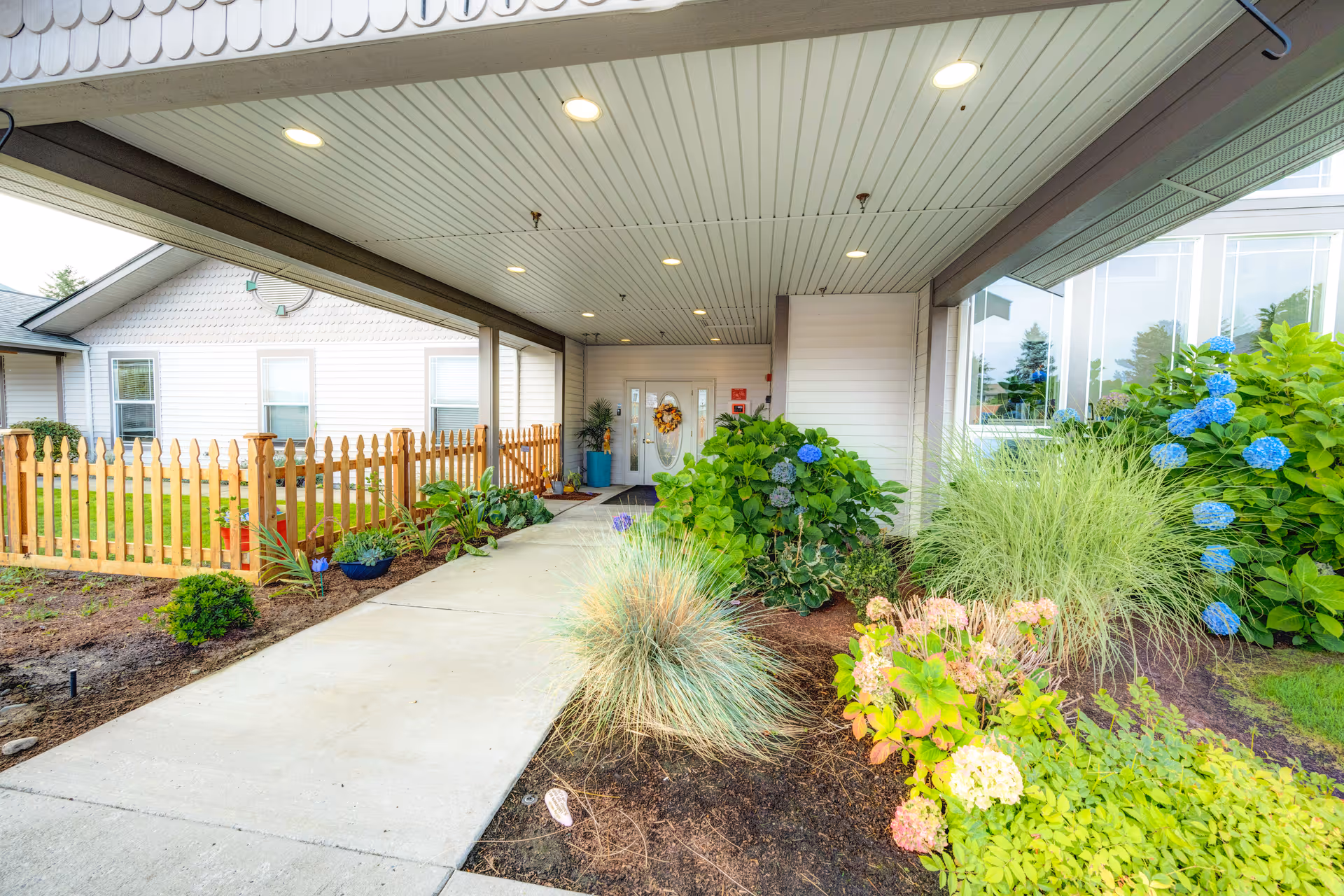 Covered front entrance walkway to a senior living facility with a picket fence, landscaped flowerbeds and a door decorated with a wreath.