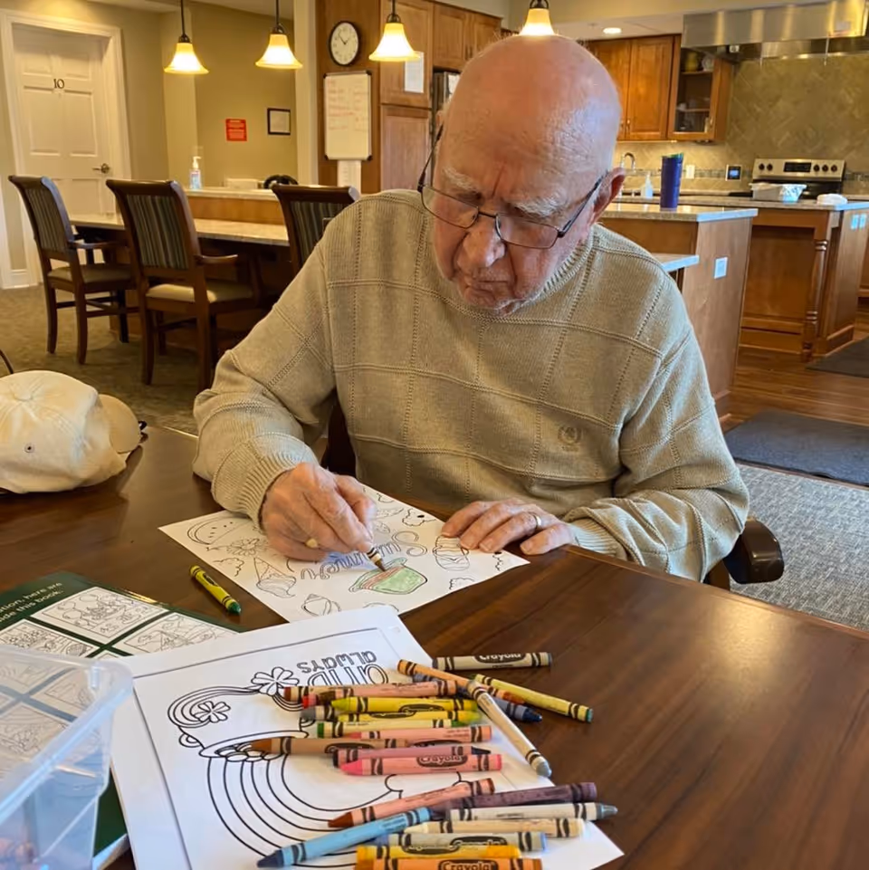 An elderly man wearing glasses and a beige sweater is sitting at a wooden table in a kitchen or dining area, coloring a picture with crayons. Various crayons and coloring pages are spread out on the table in front of him. The background shows wooden cabinets, a stove, and hanging pendant lights.