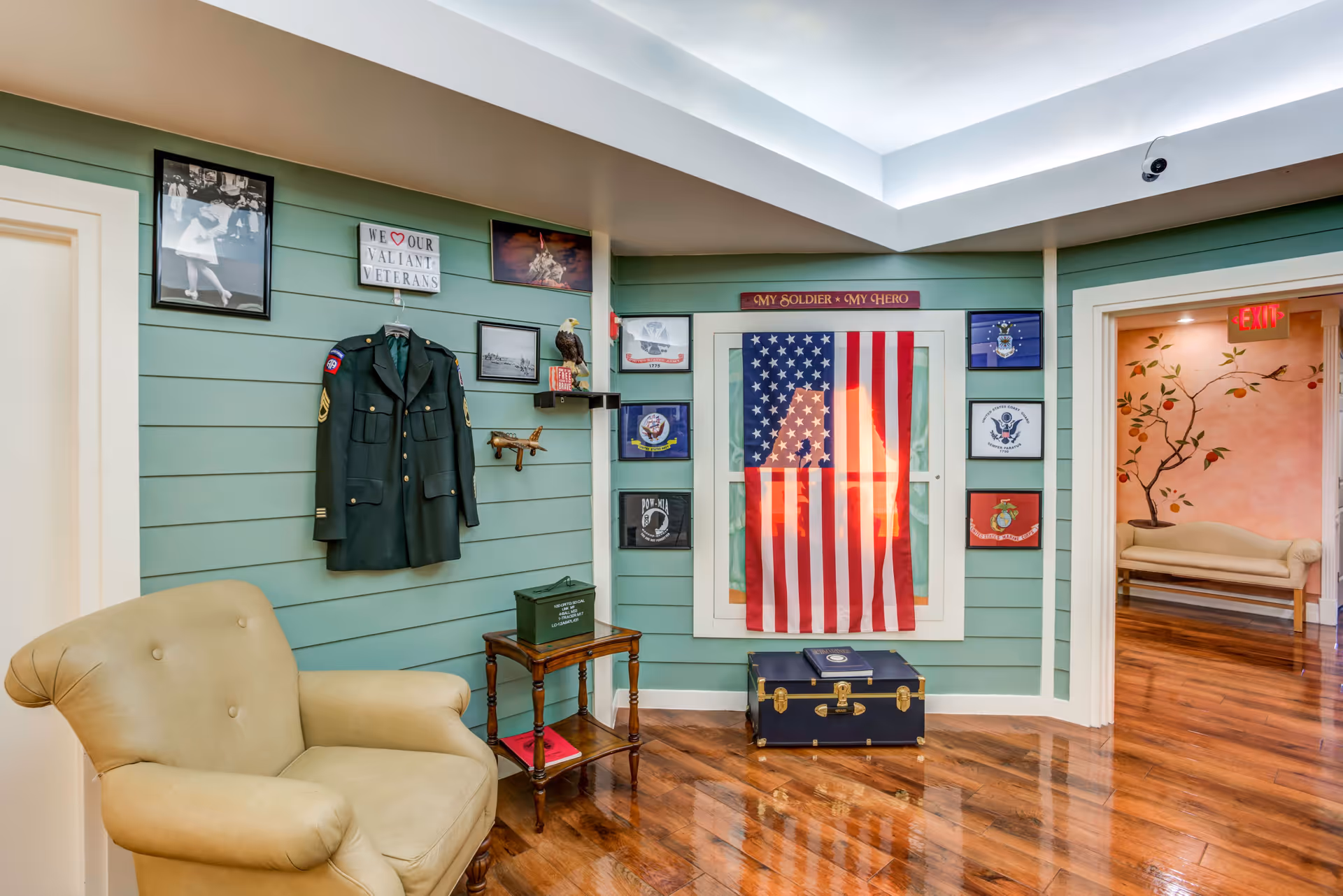 A cozy interior corner of a senior living facility with a military tribute display. The wall is painted green with horizontal panels and decorated with a military jacket, framed photos, and an American flag. A beige armchair is positioned to the left, and a small wooden table with a green box sits next to it. The floor is polished wood, and an adjacent hallway with a floral mural and a bench is visible.