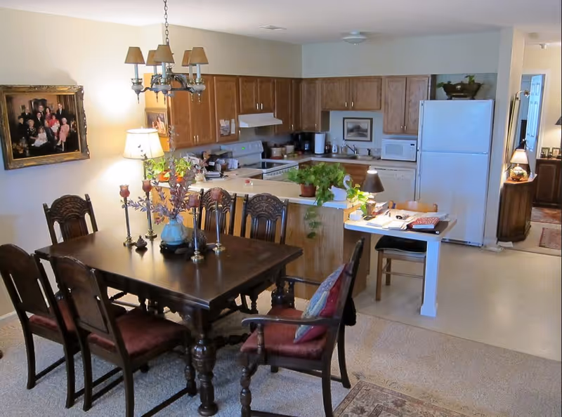 Interior view of a senior living facility showing a dining area with a dark wooden table and six chairs, decorated with a vase of flowers and candlesticks. Behind the dining area is a kitchen with wooden cabinets, a white refrigerator, microwave, stove, and countertop with potted plants. A small desk with a chair and lamp is adjacent to the kitchen. The walls are light-colored, and there are framed pictures and lamps providing warm lighting.