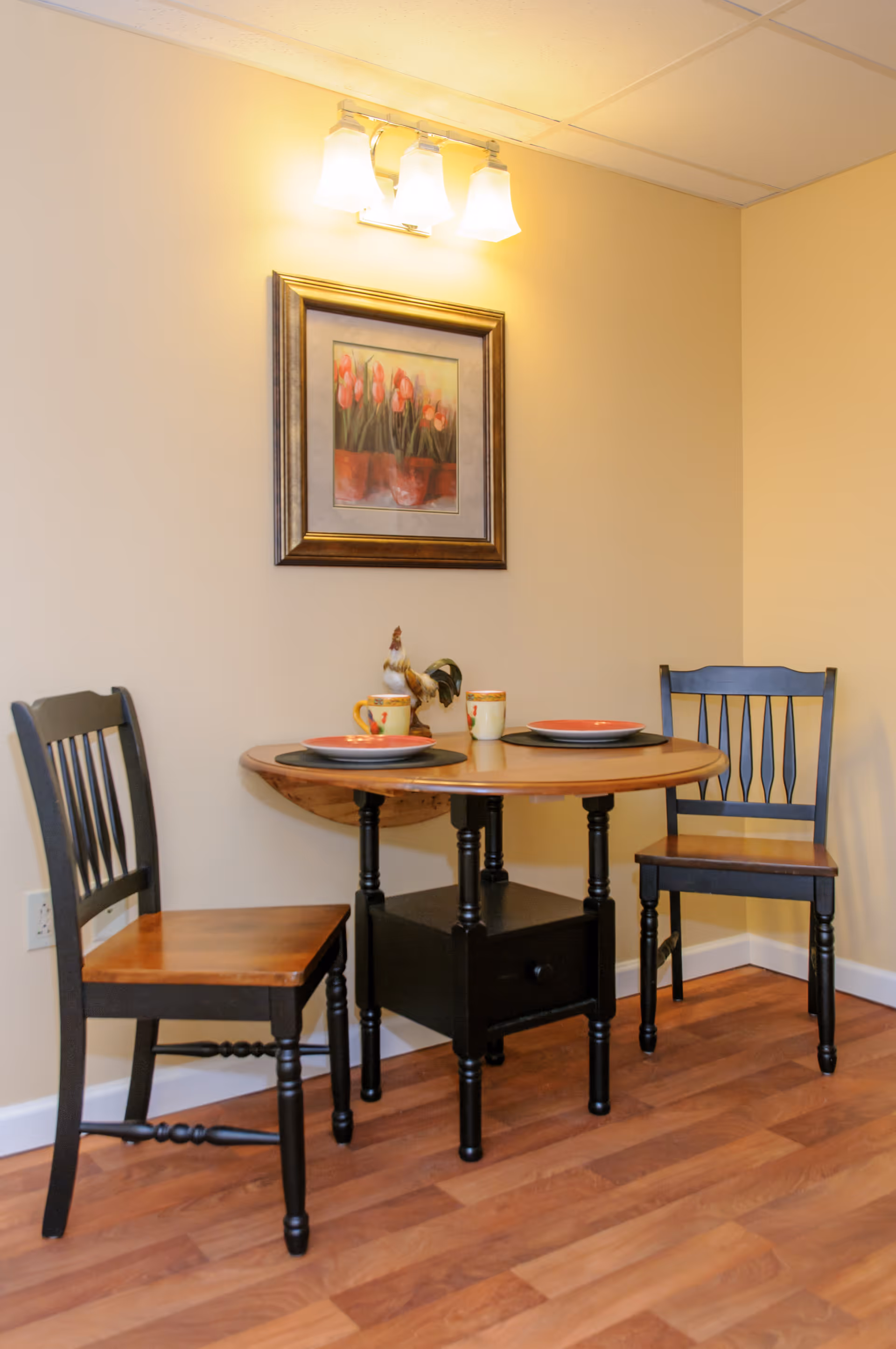 A small dining area with a round wooden table set for two, featuring two black wooden chairs with wooden seats. On the table are two plates, two mugs, and a decorative rooster figurine. Above the table is a framed painting of red tulips and a three-light wall fixture. The room has light yellow walls and wood flooring.