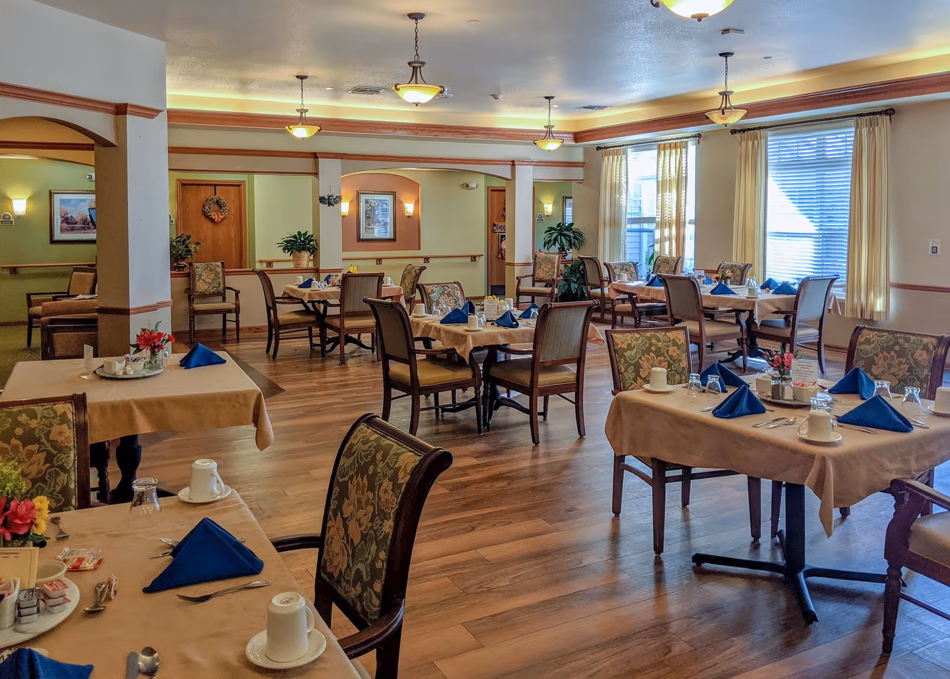 A well-lit dining room in a senior living facility with multiple tables covered in beige tablecloths, each set with blue folded napkins, white cups, glasses, and silverware. The room features wooden flooring, floral upholstered chairs, large windows with cream curtains, and decorative plants and wall art.