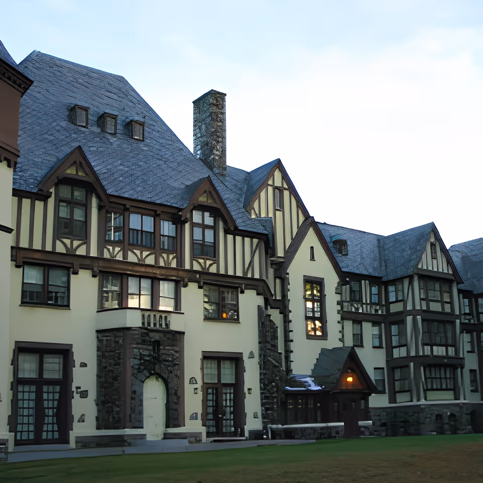 Exterior view of a large Tudor-style building with steeply pitched slate roofs, multiple windows, and stone and timber detailing. The building is set against a clear sky with a grassy lawn in the foreground.