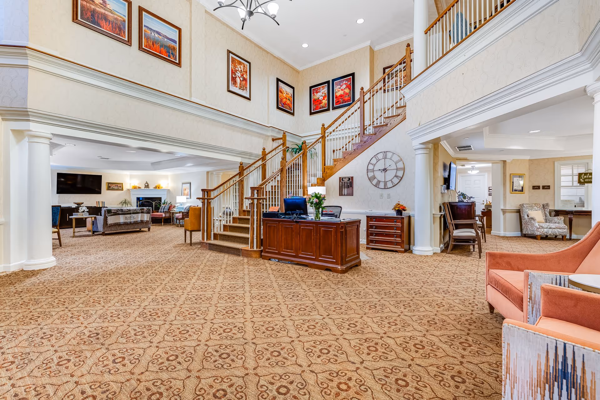 Spacious senior living facility interior with a patterned carpet, a wooden staircase with white railings, a reception desk with a computer and flowers, and a large wall clock. The room has beige walls adorned with framed artwork and a seating area with chairs and a television in the background.
