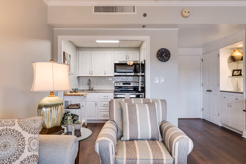 View of a cozy living room area with a striped armchair and a side table with a lamp and small plants. In the background, there is a kitchen with white cabinets, a stove, microwave, and a tiled backsplash. A clock is mounted on the wall near a white door and built-in shelves with decorative items.