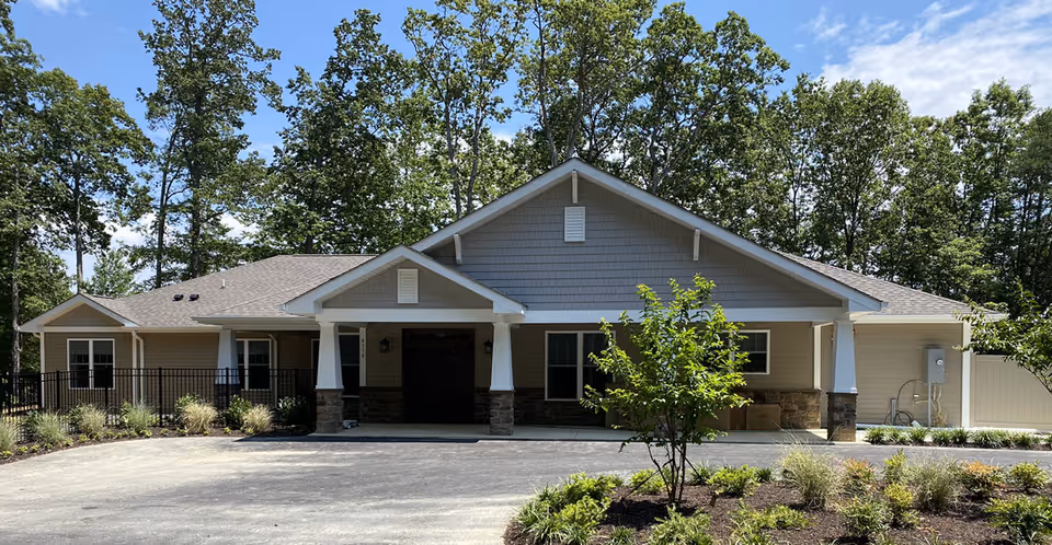 Front exterior view of a single-story senior living facility building with beige siding, stone accents, and a covered entrance supported by white columns. The building is surrounded by landscaped greenery and trees under a partly cloudy blue sky.