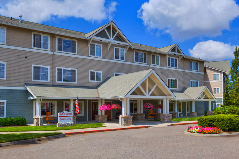 Exterior view of a three-story senior living facility building with beige siding and multiple windows. The entrance features a covered porch with stone pillars, hanging flower baskets, and benches. There is a sign near the entrance that reads 'Arbor Village Assisted Living' and an American flag displayed. The sky is blue with some clouds, and there are well-maintained shrubs and a flower bed near the driveway.