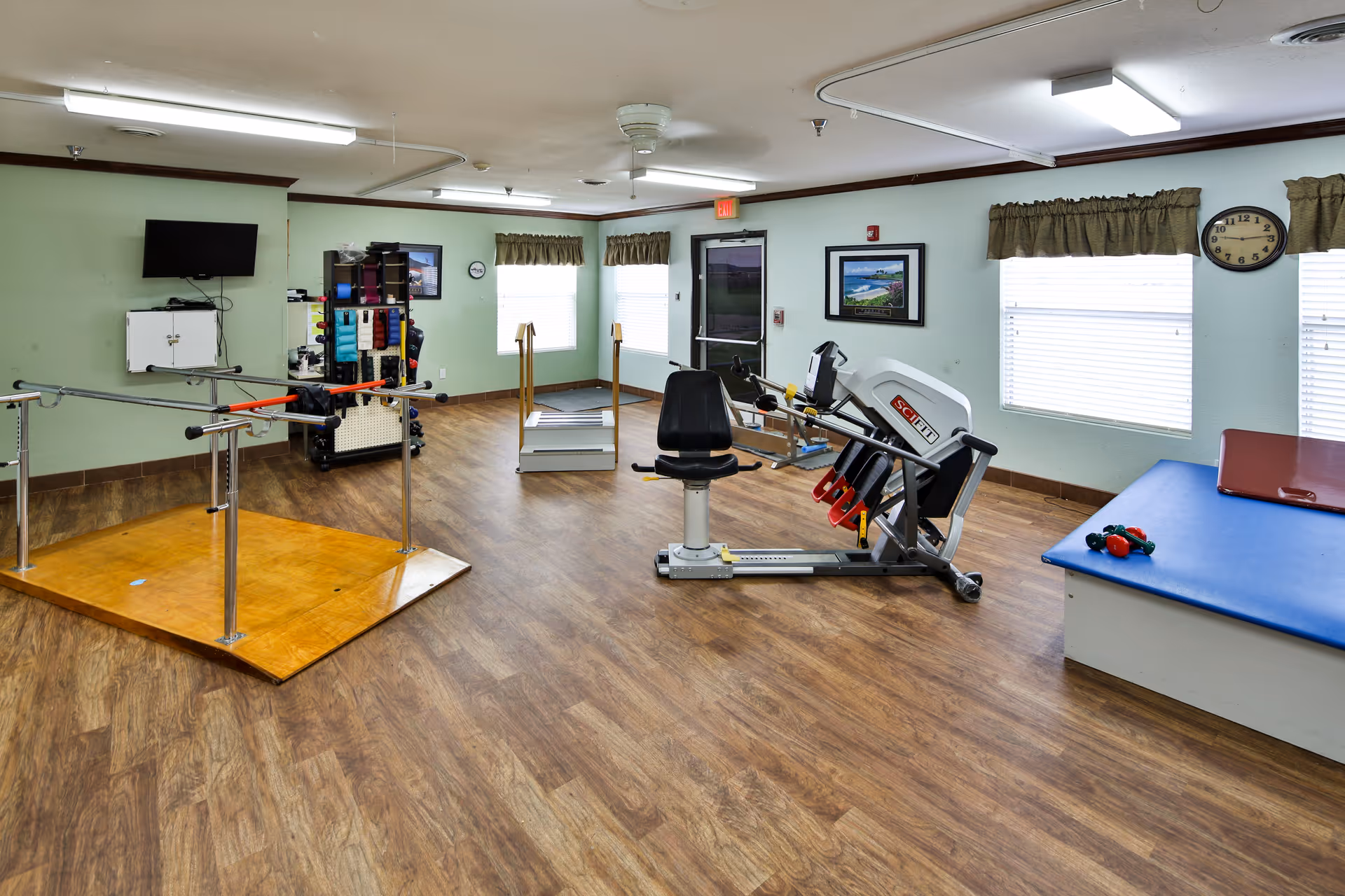 A rehabilitation room with wooden flooring and light green walls, featuring parallel bars for walking exercises, a seated leg press machine, a blue padded therapy table with small weights on it, a rack with various exercise equipment, and windows with curtains letting in natural light.