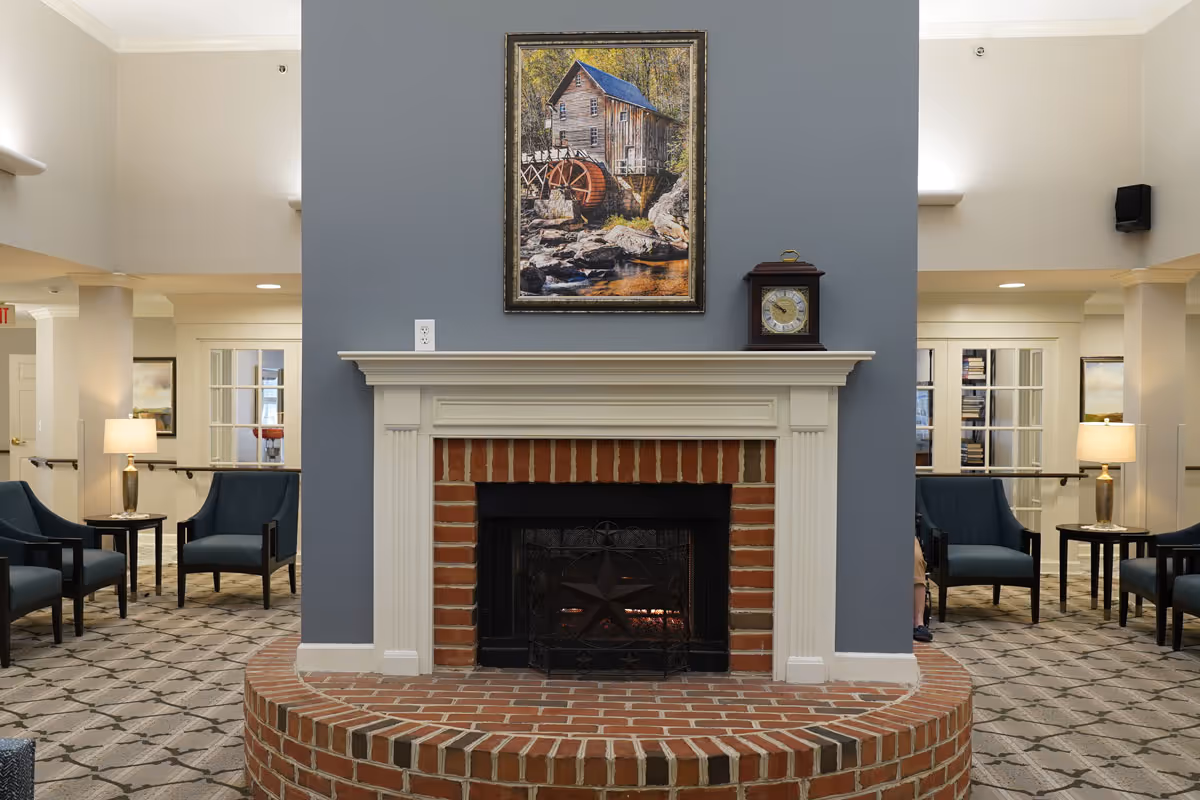 A cozy living area featuring a brick fireplace with a white mantel, above which hangs a framed painting of a watermill. On the mantel sits a vintage clock. The room has patterned carpet flooring and is furnished with blue armchairs and side tables with lamps. The walls are painted in neutral tones with a blue accent wall behind the fireplace.