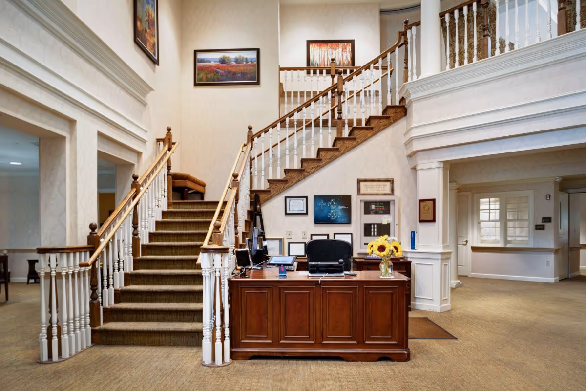 Reception area with a wooden desk and office chair in front of a staircase with wooden handrails and white balusters. The walls are decorated with framed artwork, and a vase with sunflowers is placed on the desk. The space has beige carpeting and cream-colored walls with white trim.