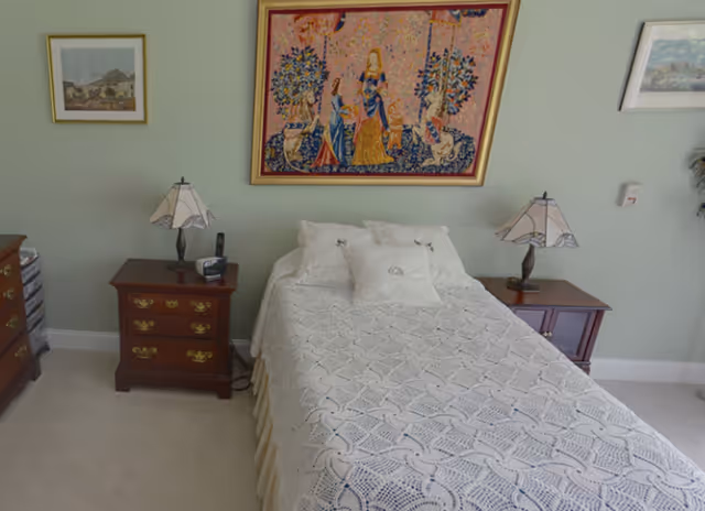 A bedroom with a single bed covered in a white crocheted bedspread and three white pillows. On either side of the bed are wooden nightstands with decorative lamps. Above the bed hangs a large framed tapestry depicting a medieval scene with three women and two animals. The walls are painted light green and have two framed pictures on either side of the tapestry.