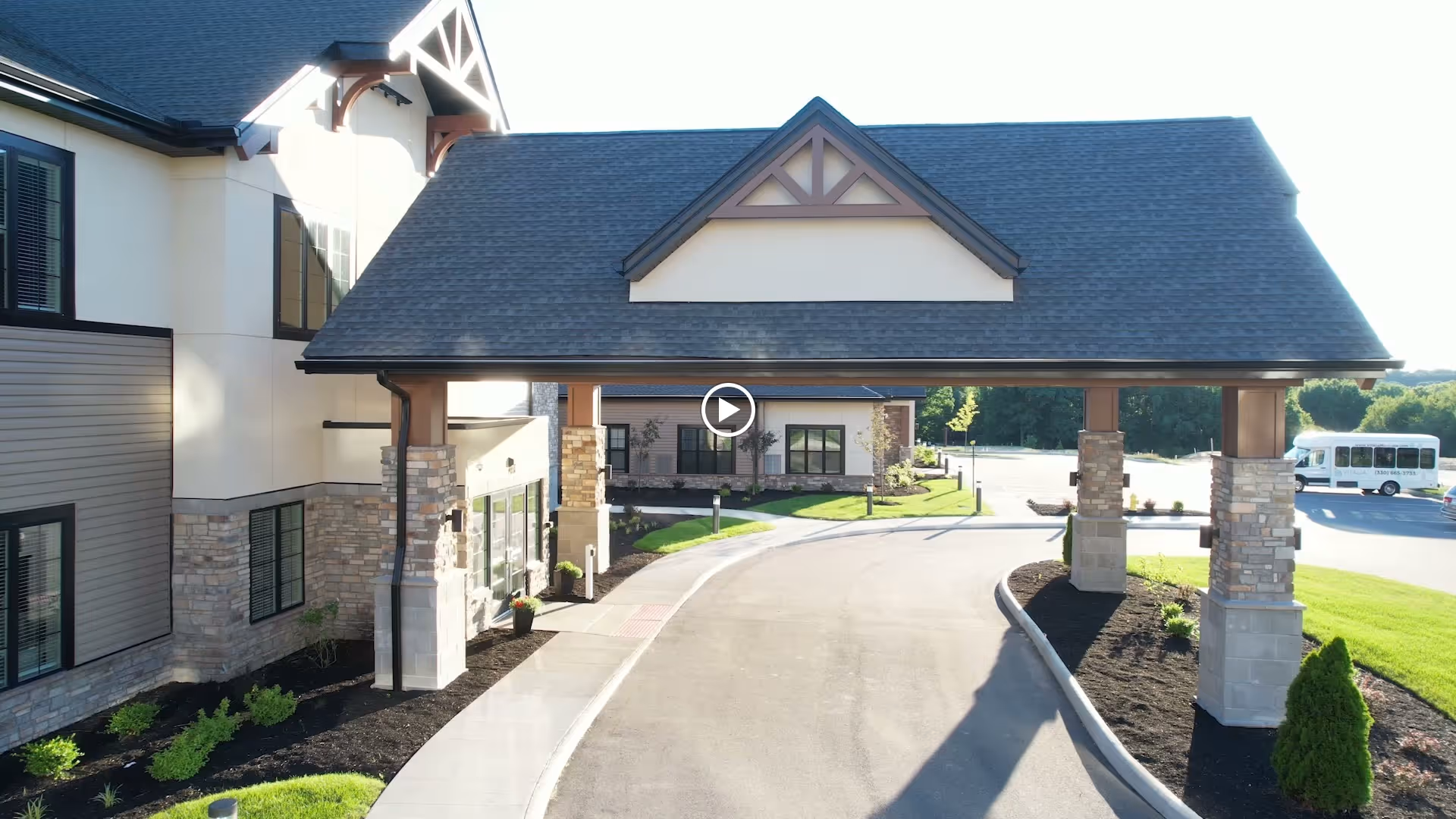 Covered entrance porte-cochere and driveway of a multi-story senior living community building with landscaping and a shuttle bus in the background.