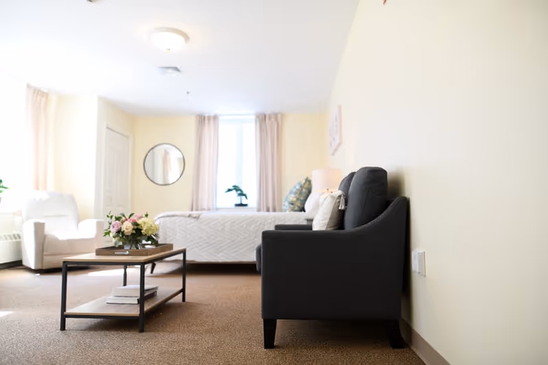A bright and cozy room with a bed near a window with beige curtains, a dark gray armchair with pillows against a light yellow wall, a white armchair near the window, and a wooden coffee table with a flower arrangement and books on it.