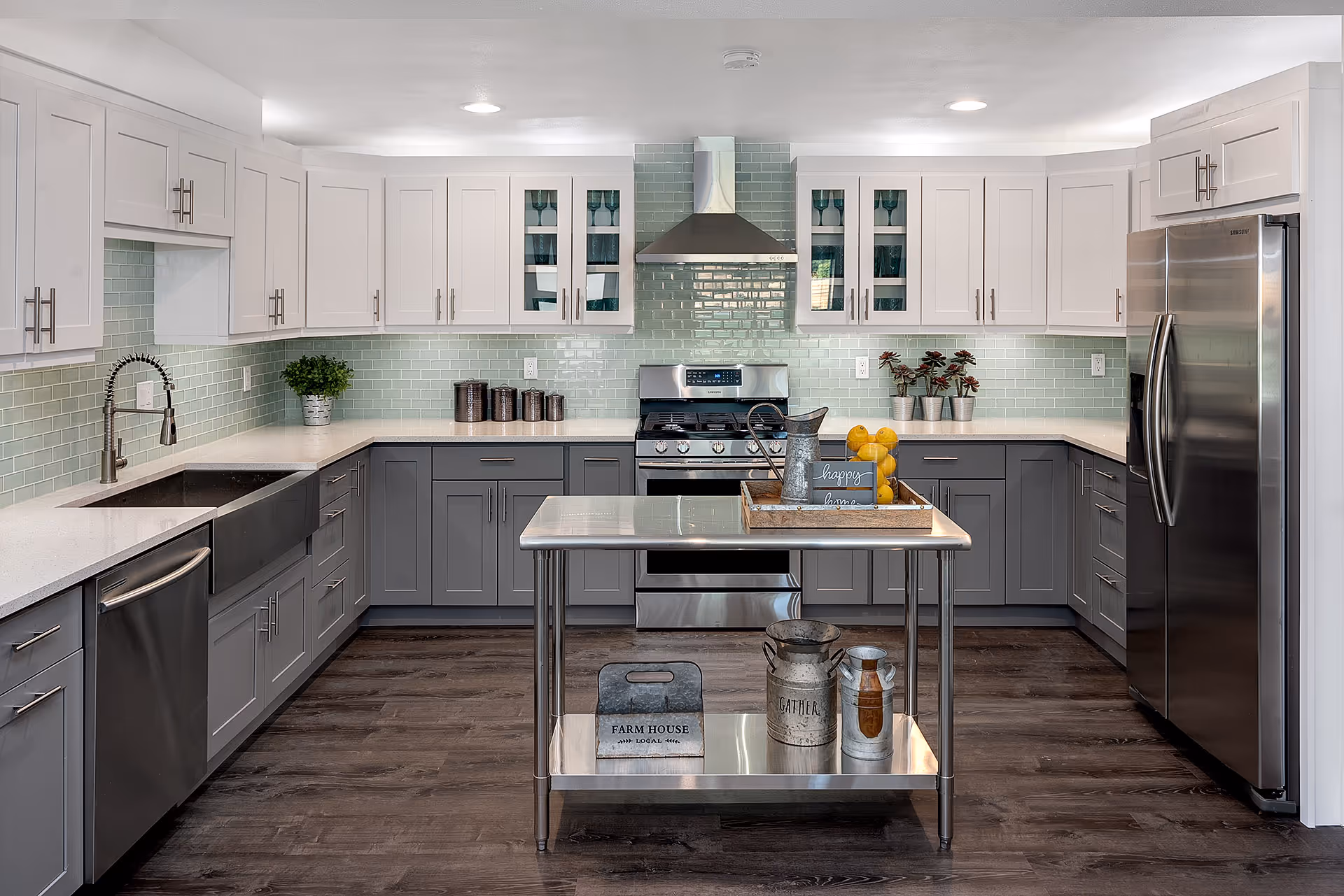 Modern kitchen with white upper cabinets and gray lower cabinets, stainless steel appliances including a refrigerator, stove, and dishwasher. There is a kitchen island with decorative items such as a tray with lemons and a watering can. The backsplash is made of light green subway tiles and the floor is dark wood.